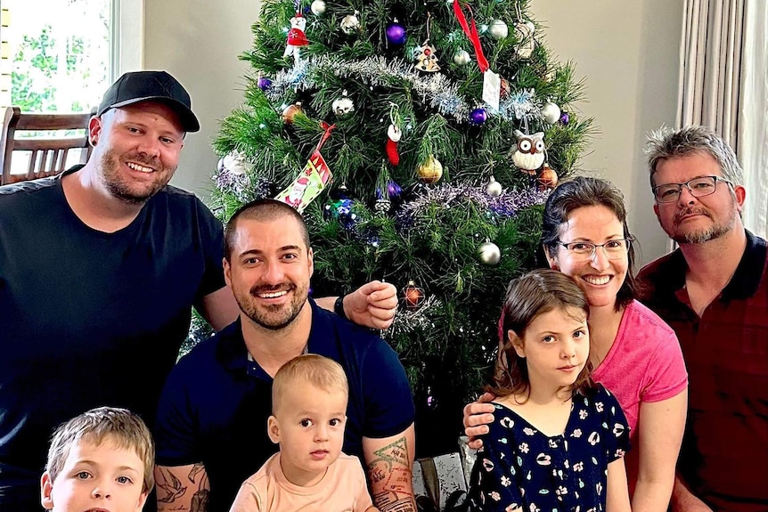 Four adults and three children pose for a photo, smiling, in front of a decorated Christmas tree