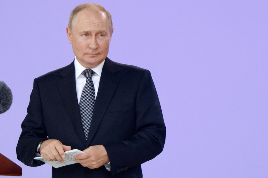 Vladimir Putin stands on a stage wearing a suit and holding some notes.