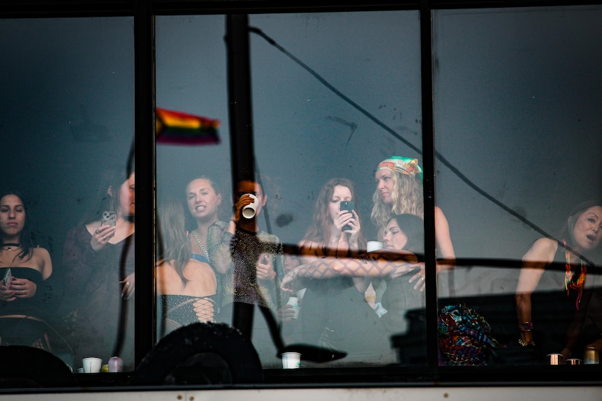 A group of young women stand behind a large sheet of glass reflecting a rainbow flag