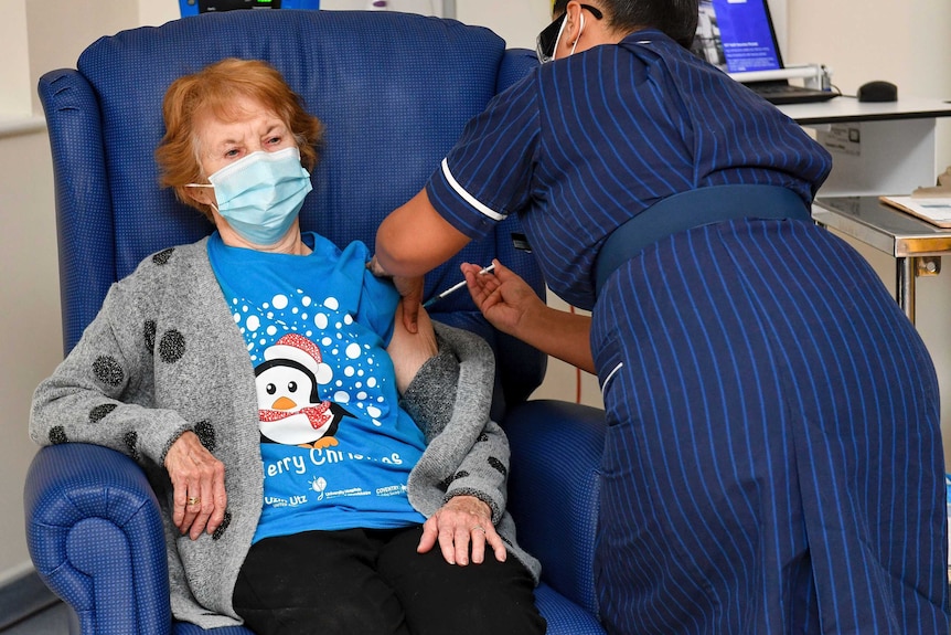 An elderly woman in a face mask sits in a chair while a nurse injects her with a needle