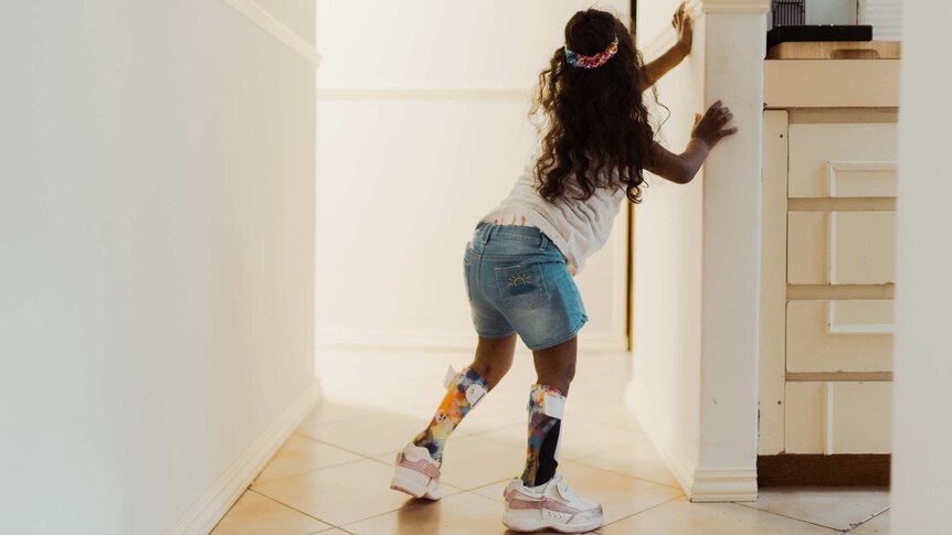 Symmie stands in the hallway, supporting herself on the wall.