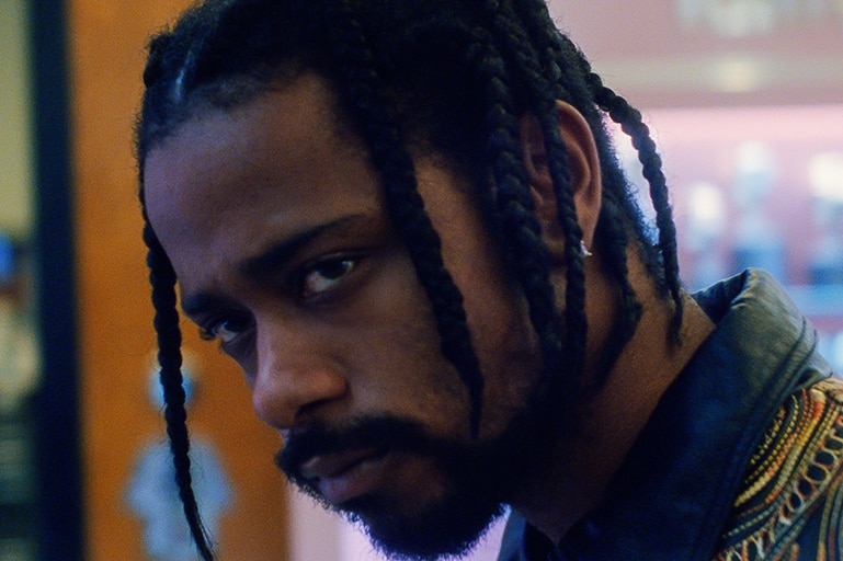 A man with black beard, short braided hair, patterned shirt and focused expression stands in a fluorescent lit shop.