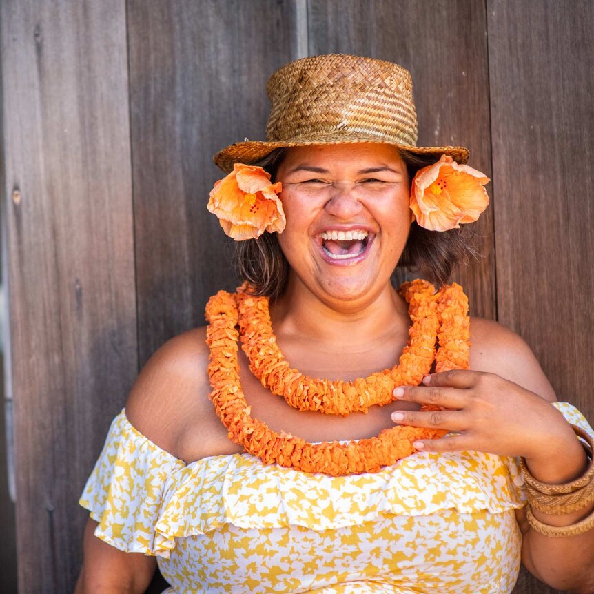 A woman in a floral dress and straw hat, with an orange lei and orange flowers in her hair laughs at the camera.
