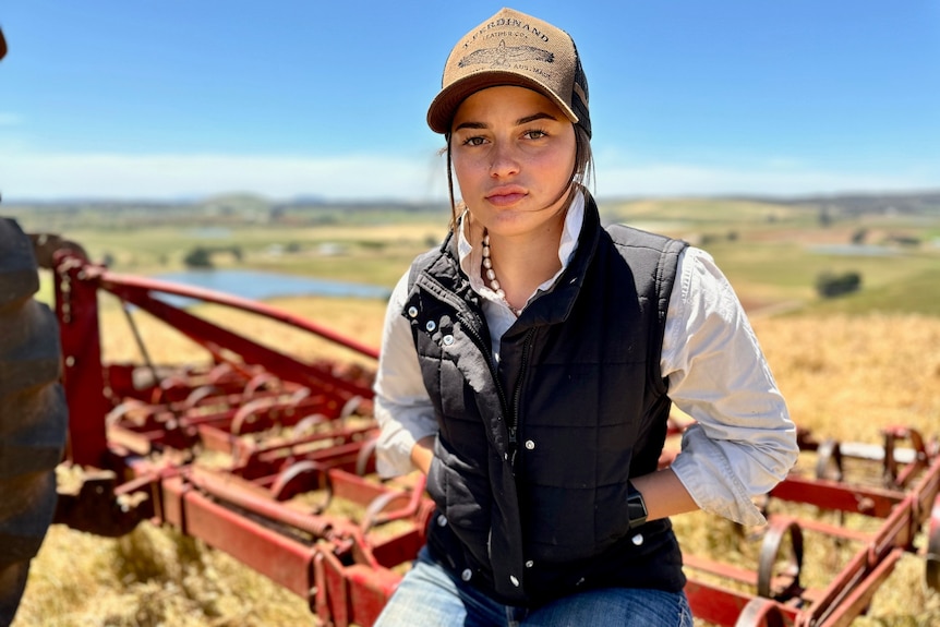 A young woman wearing a cap, black vest and work shirt with rolled up sleeves sits on a piece of machinery in a paddock.