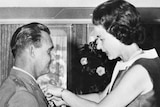 Black and white photo of soldier standing at attention in formal uniform as he has an award pinned to lapel by the Queen.