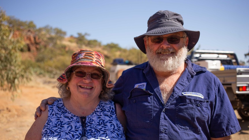 An older couple stand smiling together in the outback, with utes behind them.