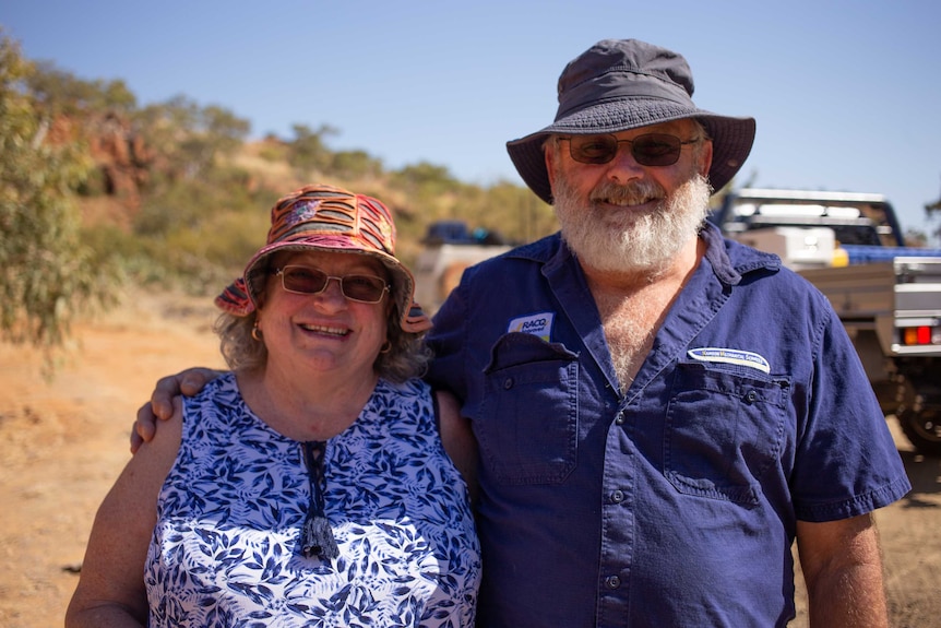 An older couple stand smiling together in the outback, with utes behind them.