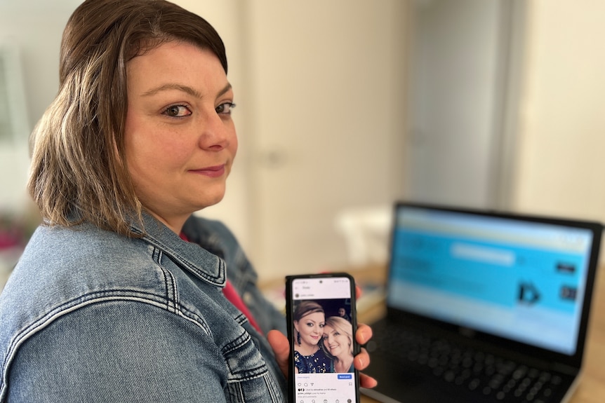 A woman with brown hair looking at the camera and holding a smartphone