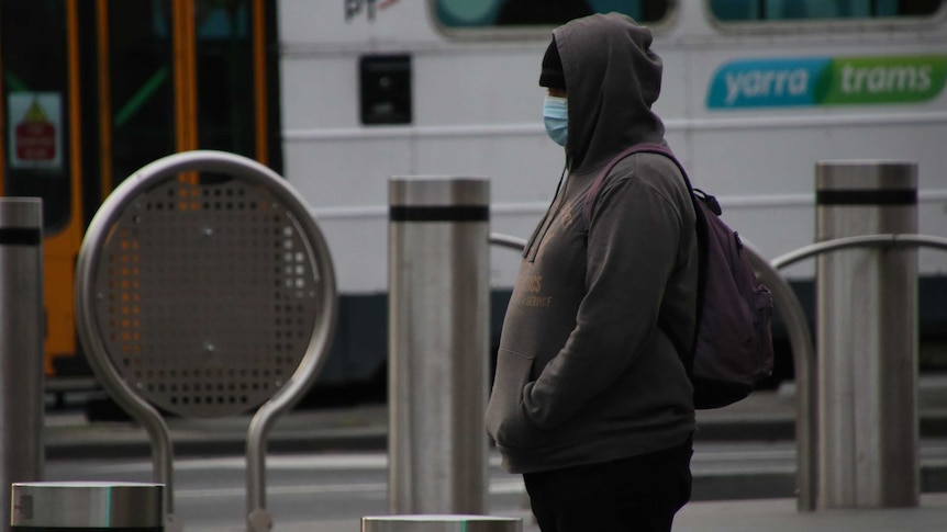 A person wearing a hood, beanie, mask stands in front of a Melbourne tram.