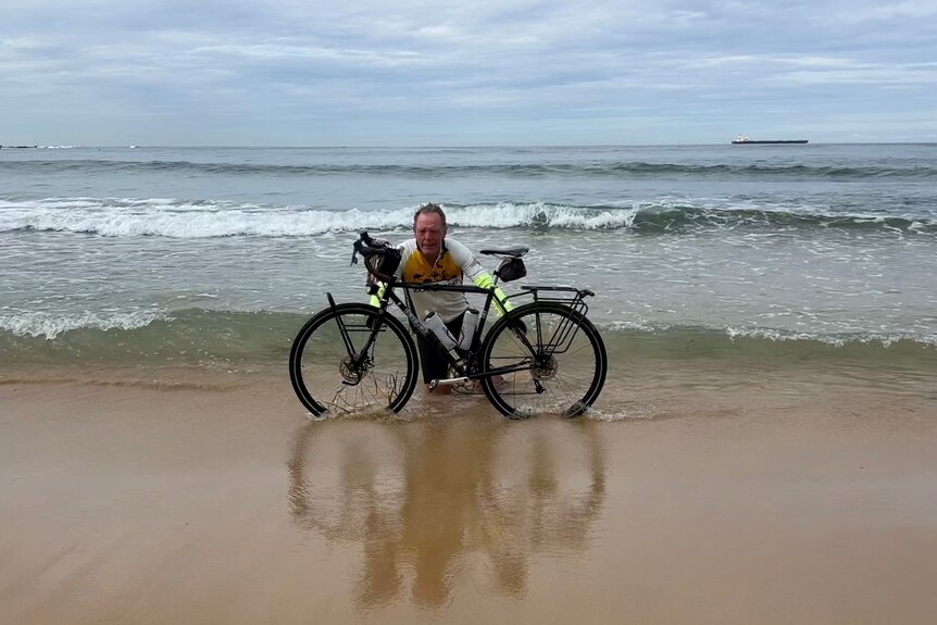 A smiling, older man in cycling gear dips the wheels of his bike in the ocean.