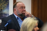 Alex Jones wears a suit and sits in a courtoom witness box, holding one hand up to his mouth