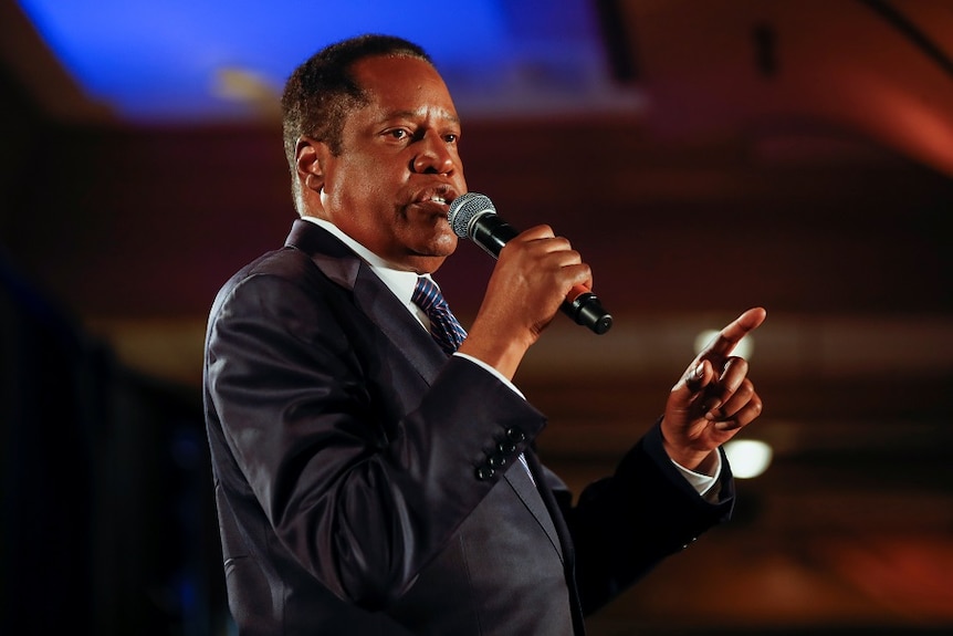 A middle-aged black man in a suit holds a microphone as he speaks at a public event.