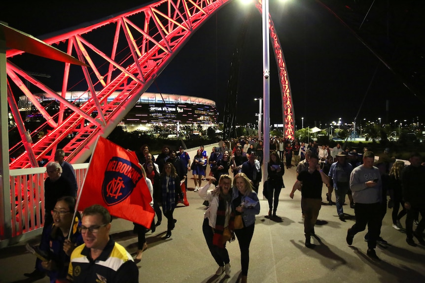 The arches above Matagarup Bridge are lit up red as fans walk across it leaving Perth Stadium at night with a Melbourne flag.