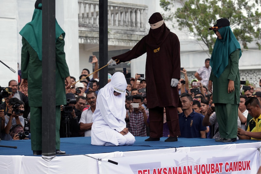 A man stands with a cane over a woman who is on her knees in on a stage in front of a crowd. 