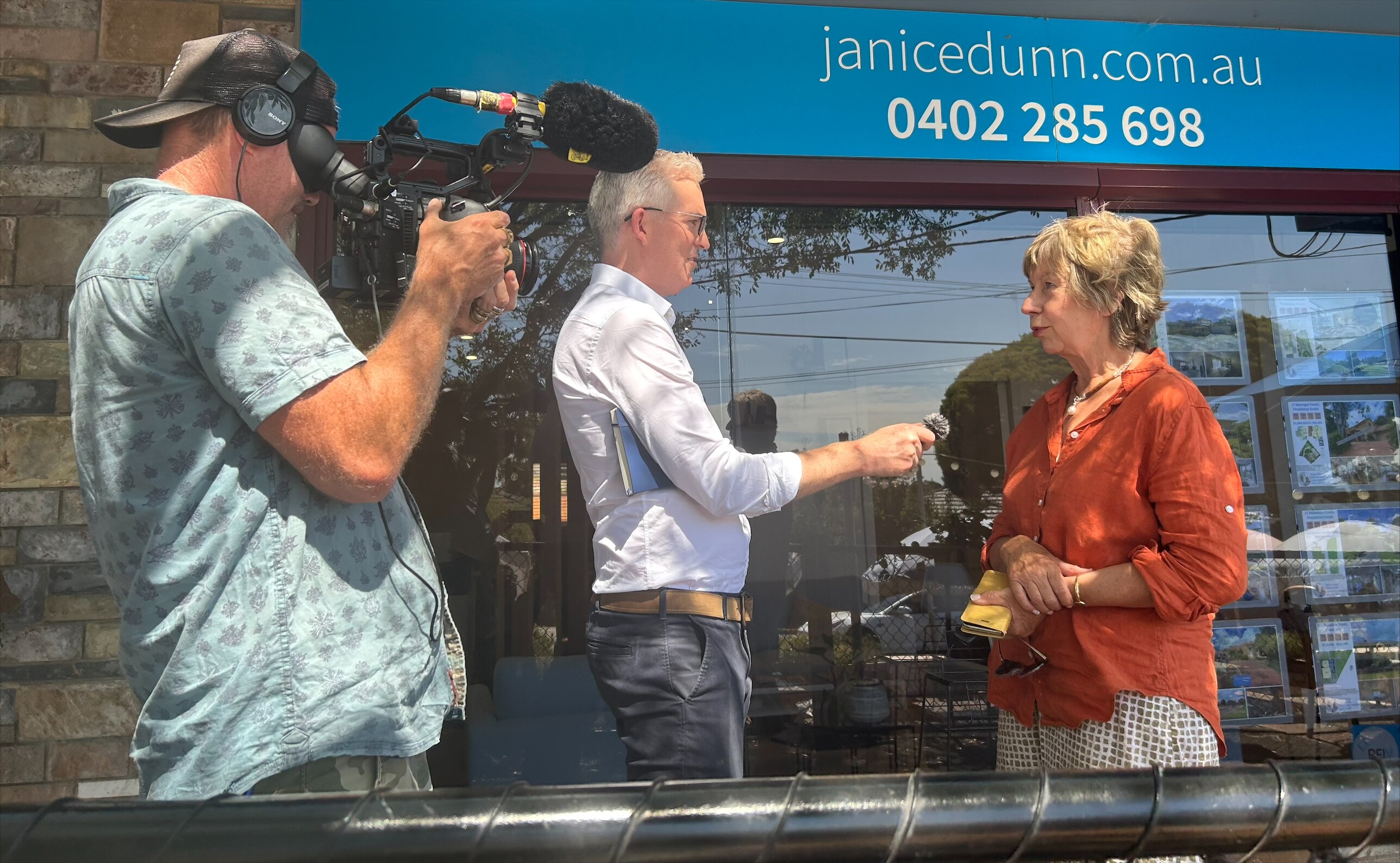 A man in a white shirt points a microphone at a woman in an orange top and a man holding a camera stands next to them 