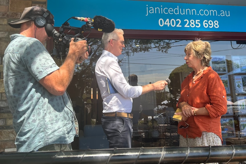 A man in a white shirt points a microphone at a woman in an orange top and a man holding a camera stands next to them 