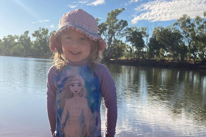 Cleo Smith stands in front of an area of water posing for a photo and smiling while wearing a hat and a Frozen shirt.