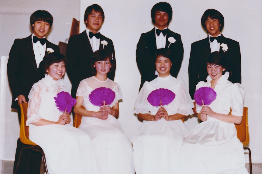 Four couples in formal attire pose together, with the women sitting in white dresses.