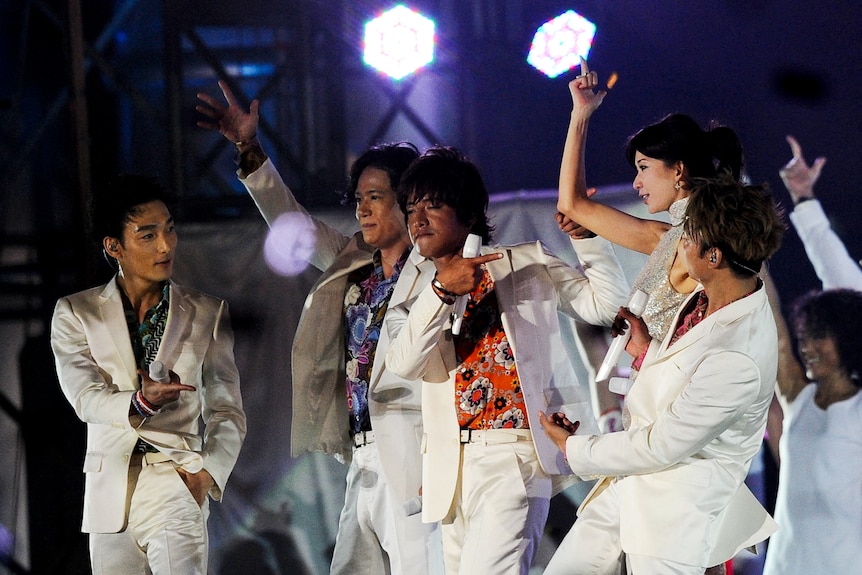 A boy band wearing white suits over garish shirts strike a pose on stage