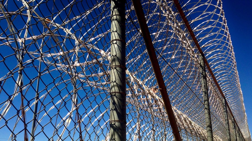 A tall fence with razor wire attached at the bottom and top. Blue sky and some jail buildings in the background.