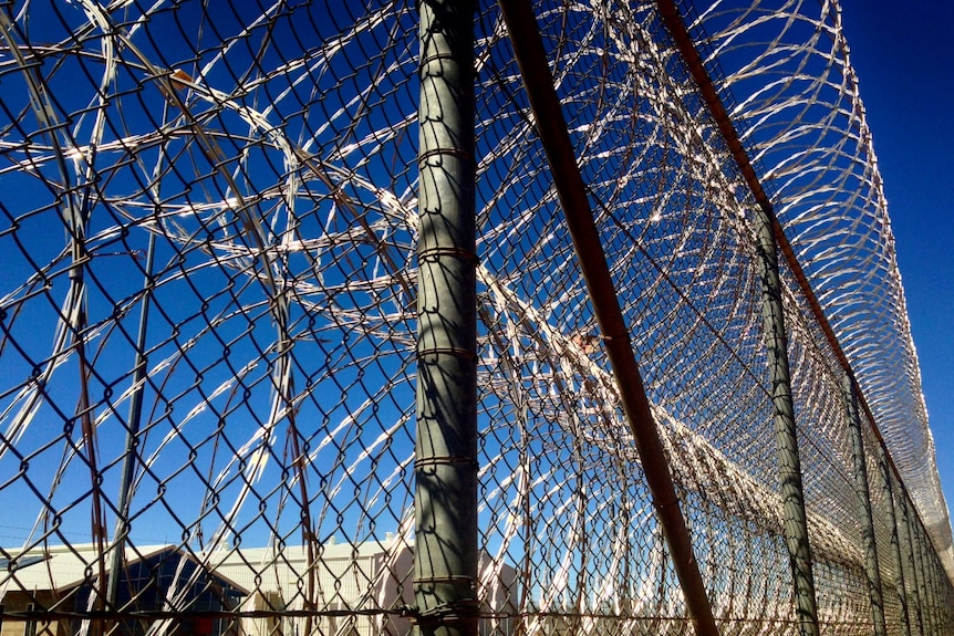 A tall fence with razor wire attached at the bottom and top. Blue sky and some jail buildings in the background.