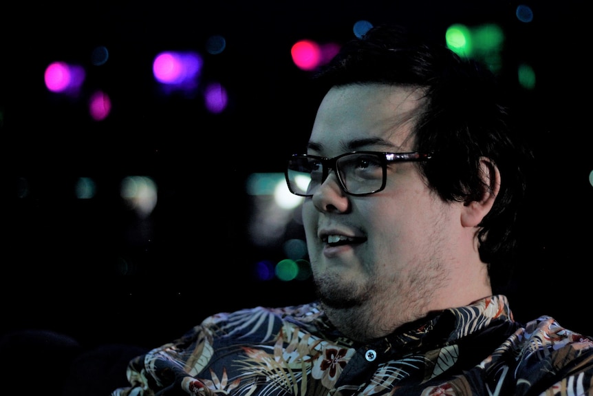 A young man in a button up shirt smiles with neon lights in the background.