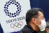Two men in masks walk past a sign reading Tokyo 2020.