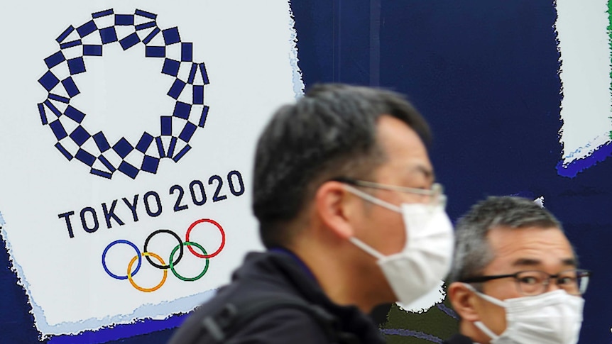 Two men in masks walk past a sign reading Tokyo 2020.