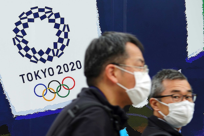 Two men in masks walk past a sign reading Tokyo 2020.