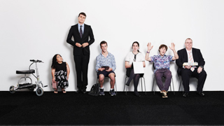 Six people stand and sit against a white wall