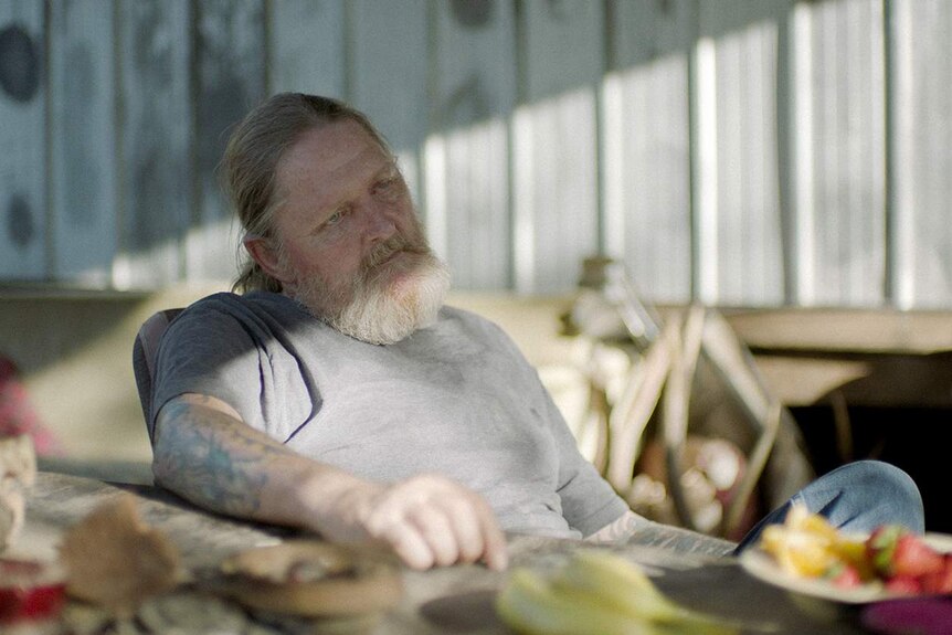 The actor, with a white beard and a whispy ponytail, sits back in a chair against a corrugated iron wall.