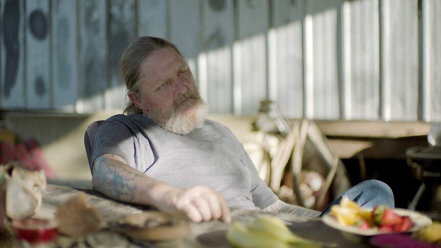 The actor, with a white beard and a whispy ponytail, sits back in a chair against a corrugated iron wall.