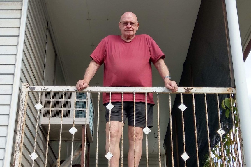 An older man wearing shorts and a tshirt and glasses looks down over a rusted balcony