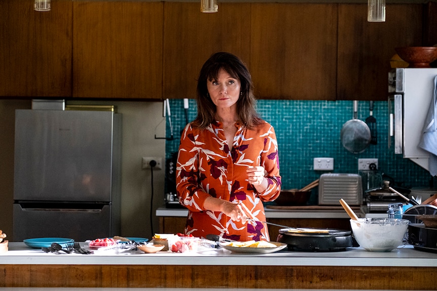 A dark haired woman in orange shirt stands preparing crepe on kitchen bench with plates and fruit with some sunlight from right.
