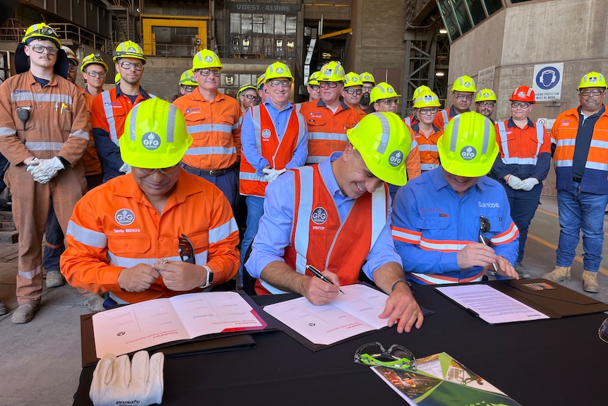 Three men in hard hats and hi-vi vest signing documents at a table as mining workers watched on