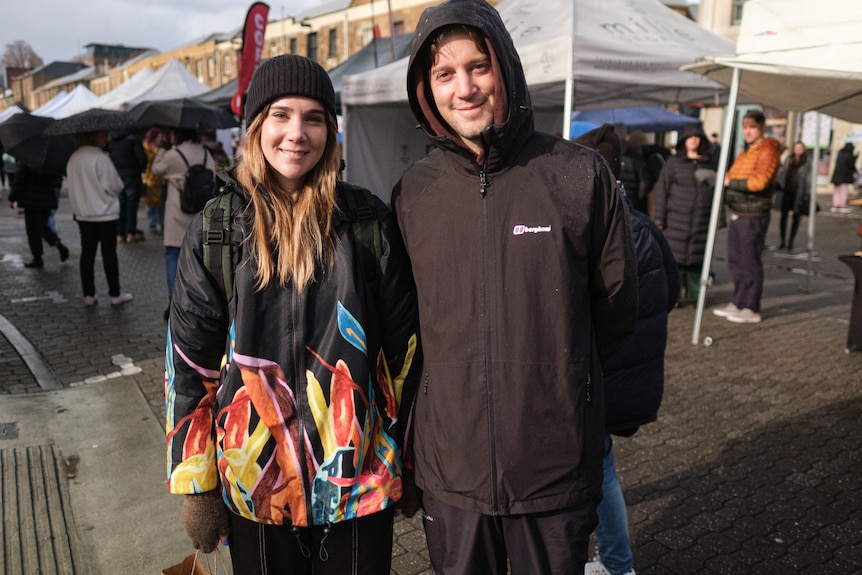 A young woman and a man in a hoodie stand at an outdoor market