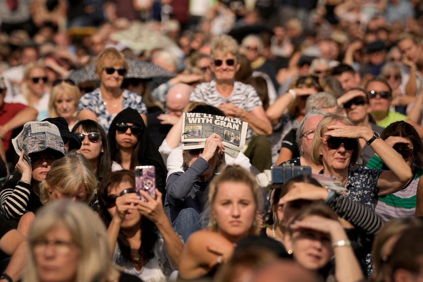 Crowds of people look on wearing sunglasses and using newspapers for shade. 