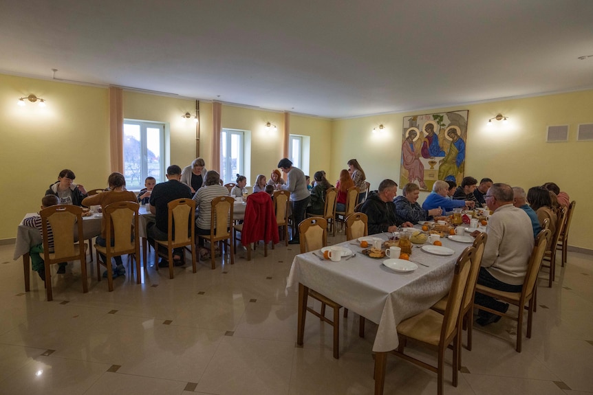 Adults and children sit around two large tables in a dining room. A religious painting is on one of the walls.