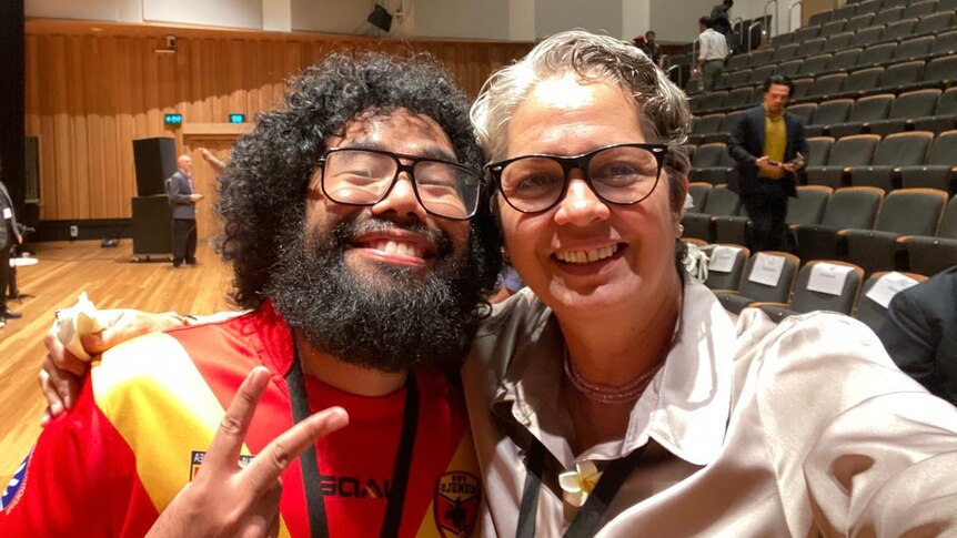A selfie of a man with curly black hair and a woman with grey hair - both wearing glasses, smiling at the camera. 