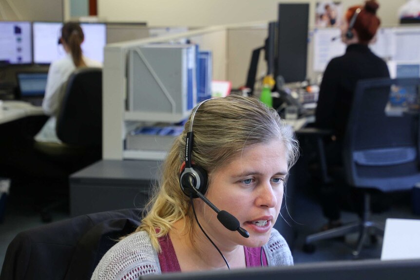 A woman sits at an office cubicle speaking on a phone headset, with other workers in the background.