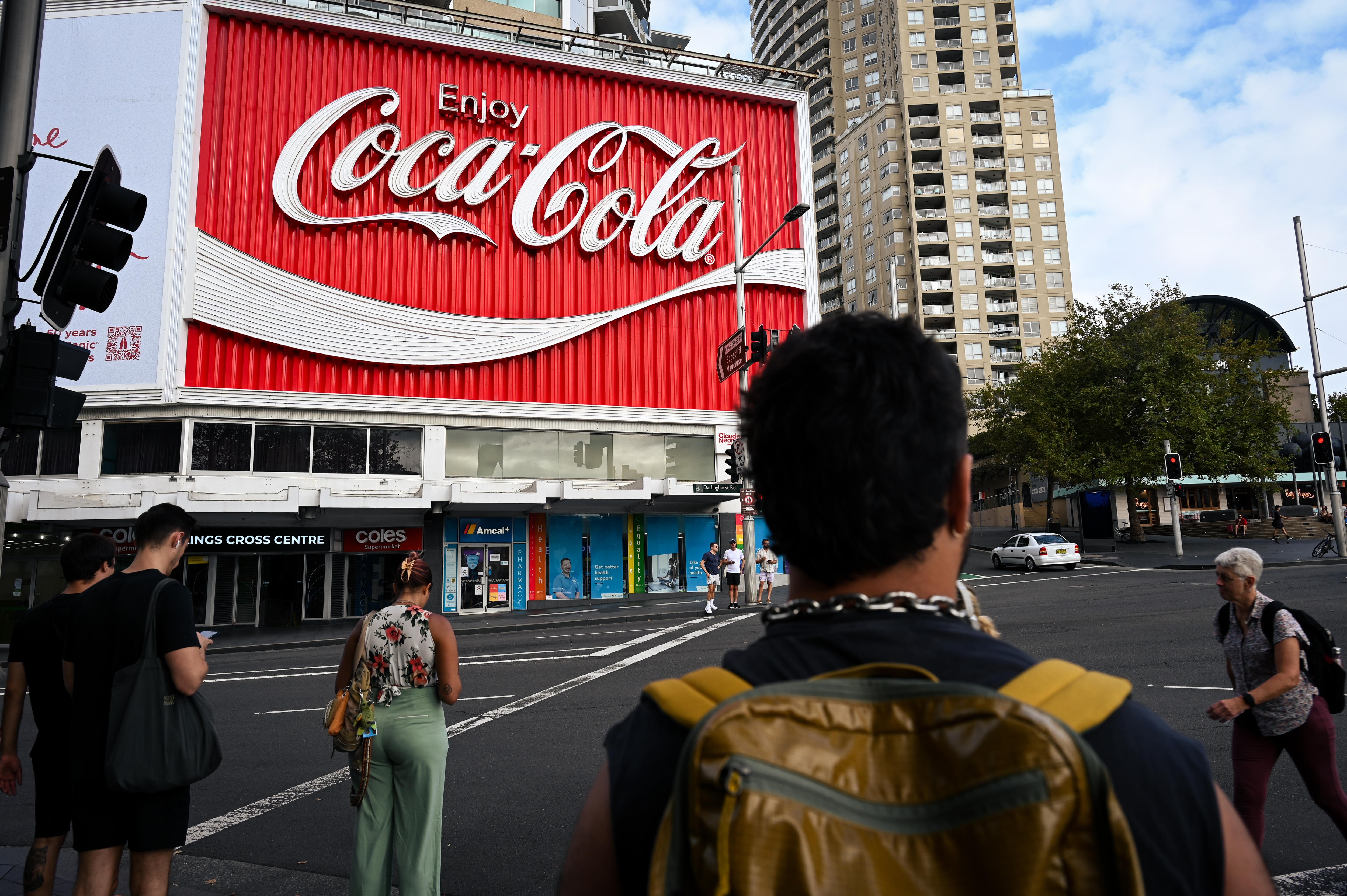The back of man wearing a yellow backpack with black hair stands in front of a coco-cola.