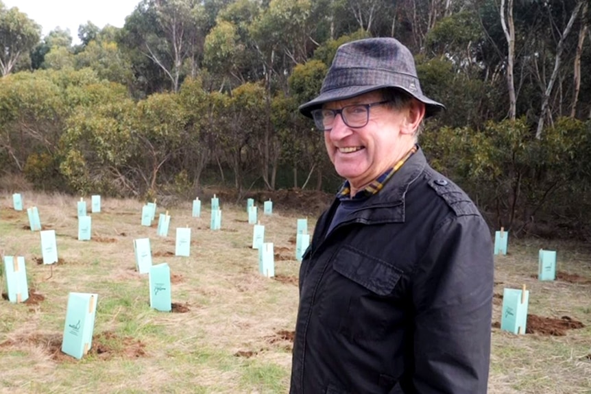 Man on right in hat, smiling looking at camera, trees in background and seedlings with green protector boxes.