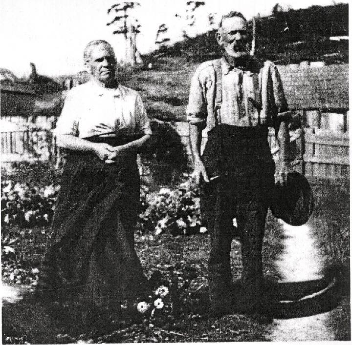 Very old black and white image of couple in front of rustic cottage.