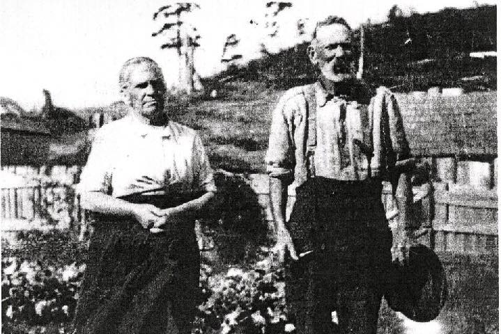 Very old black and white image of couple in front of rustic cottage.