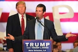 Donald Trump Jr speaks at a lectern with his hands spread out. His father looks on from behind. The US flag is in the background