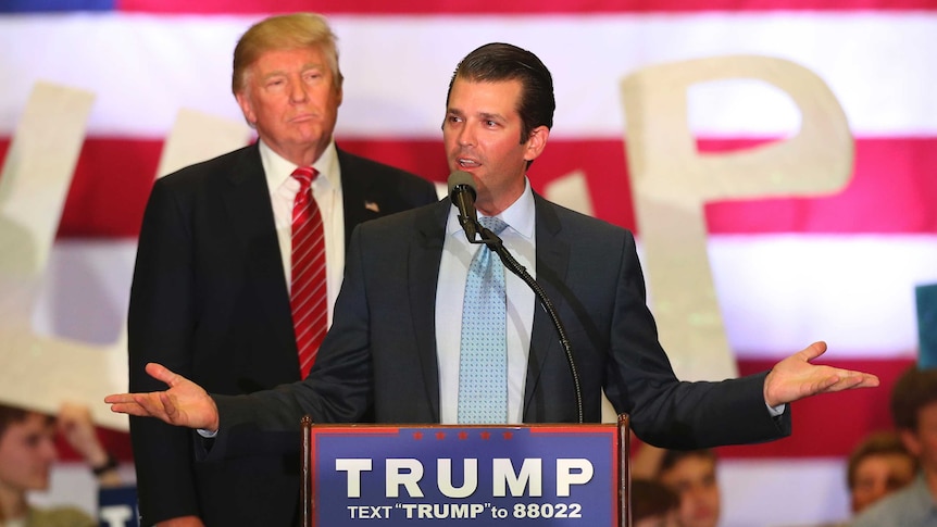 Donald Trump Jr speaks at a lectern with his hands spread out. His father looks on from behind. The US flag is in the background