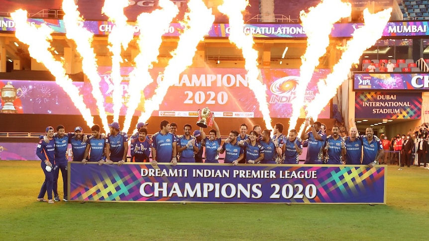 A cricket team celebrates with the IPL trophy as flames go off in the background.