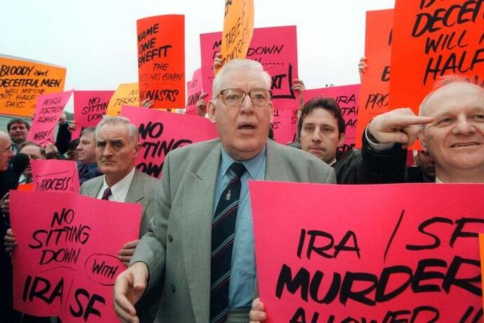 Reverend Ian Paisley stands with protesters holding bright pink and orange placard reading "NO sitting down " Murder allowed"