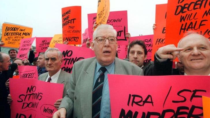 Reverend Ian Paisley stands with protesters holding bright pink and orange placard reading "NO sitting down " Murder allowed"