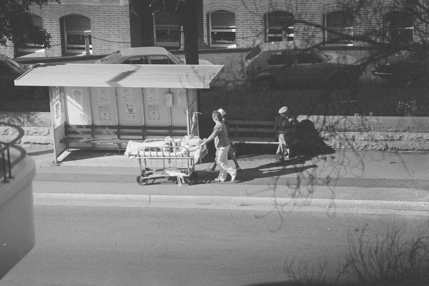 A pair of hospital workers wheel a stretcher on a footpath past a bus shelter.